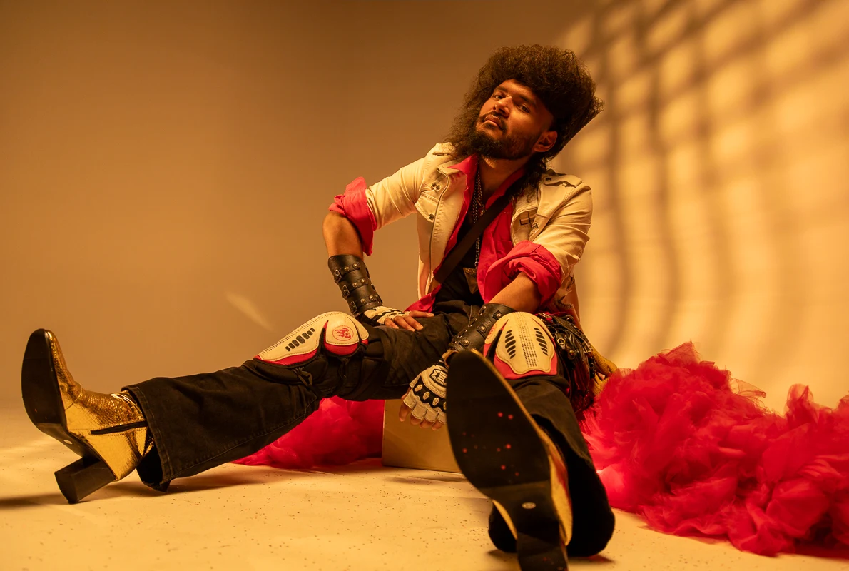 Seated performer in warm window light, with red fabric and dramatic black boots.