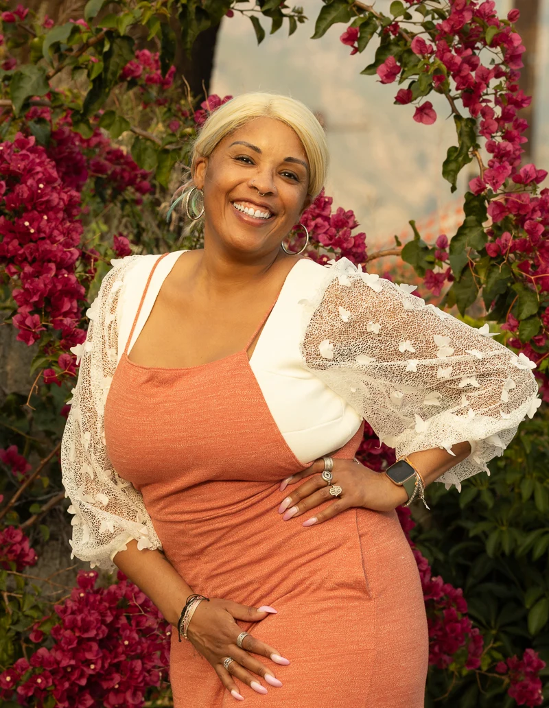 Smiling person in a peach dress and white lace jacket, surrounded by pink flowers.